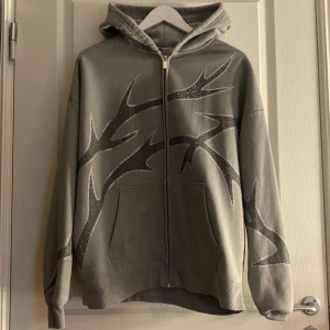 Grå hoodie med strassdetaljer från FB Sister - Säljer en grå hoodie från FB Sister med coola strassdetaljer i ett mönster över hela tröjan. Den har en dragkedja framtill och en bekväm huva. Perfekt för att ge din outfit lite extra bling! 💎Obs! Köpt för 329kr