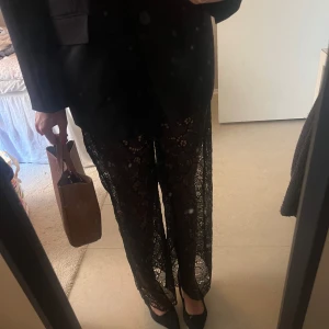 Svarta spetsbyxor - Eleganta svarta byxor i spets med vida ben och hög midja. Perfekta för en stilfull look med en touch av glamour. Kombinera med en enkel topp för att låta byxorna stå i centrum.