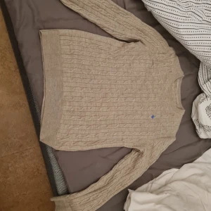 Beige stickad tröja med blå detalj - Säljer en beige stickad tröja med ett diskret blått märke på bröstet. Tröjan har ett klassiskt kabelstickat mönster och långa ärmar. Perfekt för kyligare dagar.