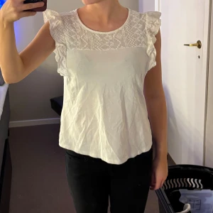 Vit blus med spetsdetaljer - Säljer en söt vit blus med spetsdetaljer på axlarna. Blusen har en rund halsringning och är ärmlös med volangdetaljer. Perfekt för en somrig look!