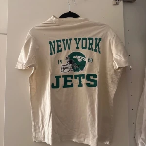 Vit New York Jets t-shirt - Säljer en vit t-shirt med New York Jets tryck i grönt. T-shirten har korta ärmar och ett stort tryck med texten 'New York Jets' och en hjälm på baksidan. Perfekt för sportfantasten!