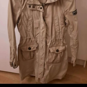 Beige parkajacka från Peak Performance - Säljer en snygg beige parkajacka från Peak Performance. Jackan har långa ärmar, dragkedja och knappar framtill samt praktiska fickor med lock. Perfekt för höst och vår med sin stilrena design och bekväma passform. 