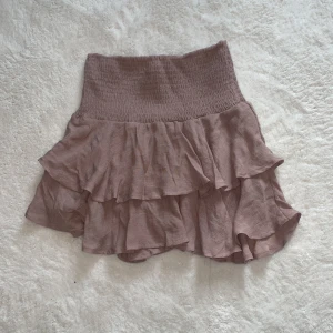 Beige volangkjol - Säljer en söt beige kortkjol med volanger. Kjolen har en elastisk midja för bekväm passform och är perfekt för varma dagar. Den luftiga designen ger en romantisk känsla. Passar bra till en enkel topp eller blus.