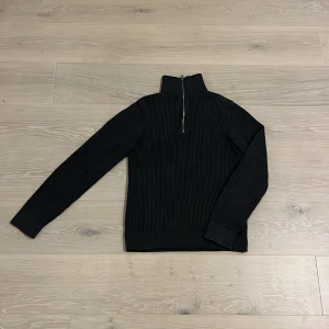 Svart ribbad tröja från East West - Säljer en stilren svart ribbad tröja från East West. Tröjan har en dragkedja framtill och en hög krage som kan vikas ner. Perfekt för kyligare dagar.