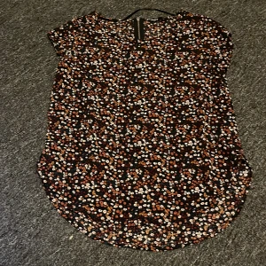 Blommig topp från ONLY - Säljer en söt blommig topp från ONLY i storlek 34. Toppen har ett färgglatt blommönster i orange, vitt och rött på en svart bakgrund. Den är kortärmad och har en rundad nederkant. Perfekt för en avslappnad stil.