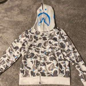 Bape tröja - Säljer en grå camo hoodie från A Bathing Ape med ett unikt hajmotiv på huvan. Tröjan har en dragkedja framtill och är långärmad. Perfekt för dig som vill sticka ut med en cool och trendig stil.