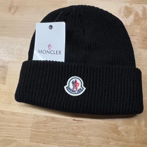 Svart ribbad mössa från Moncler - Snygg svart ribbad mössa från Moncler med broderad logga framtill. Perfekt för kyliga dagar med stil. Passar både höst och vinter.