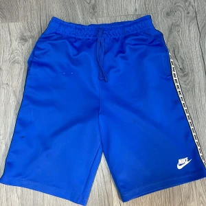 Blå shorts från Nike - Snygga blå shorts från Nike med vit logga och sidoband med Nike-text. Perfekta för träning eller en avslappnad dag. De har en elastisk midja med snörning för bästa passform.
