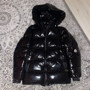 Svart glansig pufferjacka med huva - Snygg svart glansig pufferjacka från Moncler med huva och dragkedja. Perfekt för kalla dagar med sin quiltade design som håller värmen. Jackan har långa ärmar och en stilren look. Säljer den för att den passar inte mig längre.