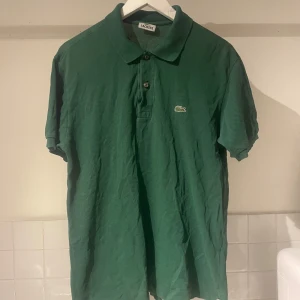 Grön pikétröja från Lacoste - Säljer en klassisk grön pikétröja från Lacoste med den ikoniska krokodilloggan på bröstet. Tröjan har korta ärmar och knappar vid halsen. Perfekt för en avslappnad stil.