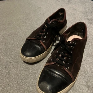 Bruna sneakers från Lanvin - Snygga bruna sneakers från Lanvin med svarta detaljer och snörning. Skorna har en klassisk design med en vit sula och är perfekta för en stilren look.