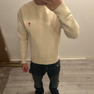 Beige tröja med röd detalj från AMI - Säljer en stilren beige tröja med en liten röd broderad detalj på bröstet. Tröjan har långa ärmar och ribbade muddar vid ärmslut och nederkant. Perfekt för en avslappnad look.