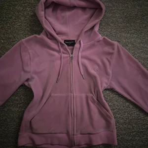 Rosa hoodie från Juicy Couture - Säljer en mysig rosa hoodie från Juicy Couture med dragkedja och huva. Perfekt för en avslappnad stil. Den har en praktisk ficka framtill och ett snyggt J-detalj på dragkedjan.