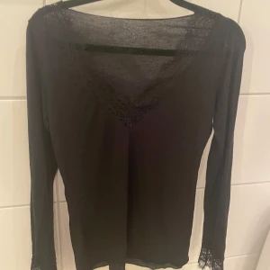 Svart långärmad topp med spetsdetaljer - Elegant svart topp med långa ärmar och v-ringad hals. Toppen har vackra spetsdetaljer vid halsringningen och ärmsluten, vilket ger en stilren och sofistikerad look. Nypris 450kr