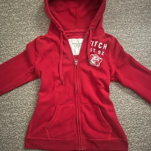 Röd hoodie från Abercrombie & Fitch - Säljer en snygg röd hoodie från Abercrombie & Fitch med dragkedja och huva. Den har ett coolt tryck med texten 'FITCH EST 92' och en broderad tiger på bröstet. Perfekt för en avslappnad stil!