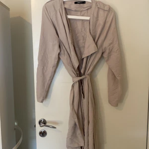 Beige trenchcoat från Gina Tricot - Säljer en stilren beige trenchcoat från Gina Tricot. Jackan har en klassisk design med långa ärmar och en avslappnad passform. Perfekt för vår och höst! Ena hålet har gått av till fästet men de går o sy tbx 