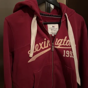 Röd hoodie från Lexington - Säljer en snygg röd hoodie från Lexington med dragkedja och vit text 'Lexington 1993' på framsidan. Den har en justerbar huva med vita snören och praktiska fickor framtill. Perfekt för en avslappnad stil.