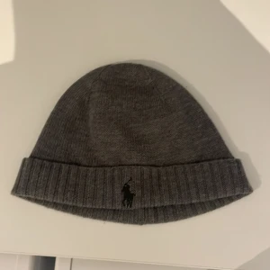 Grå stickad mössa från Ralph Lauren - Snygg grå stickad mössa från Ralph Lauren med broderad logotyp framtill. Perfekt för kyliga dagar. Säljer denna mössa för att jag hitta min gamla mössa därför har ingen användning 