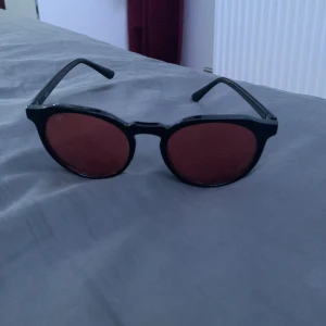 Svarta solglasögon med runda röda linser - Snygga svarta solglasögon med runda röda linser. Perfekta för att ge en cool touch till din stil. Passar både för soliga dagar och som en trendig accessoar.