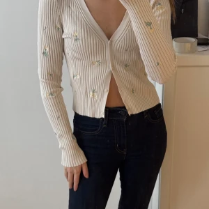 Beige ribbad kofta med blommor - Säljer en beige ribbad kofta med små broderade blommor i gult och grönt. Koftan har en v-ringad design och knappar framtill. Perfekt för en stilren och söt look.