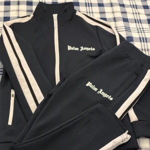 Marin blå träningsset från Palm Angels - Snyggt marin blå träningsset från Palm Angels med vita ränder längs ärmar och ben. Jackan har dragkedja och två fickor med dragkedjor. Byxorna har elastisk midja och matchande ränder. Perfekt för en sportig look.