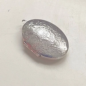 Silverfärgd ovalt medaljong - Elegant oval medaljong i silverfärg med vackert blommönster. Perfekt för att bära nära hjärtat. Medaljongen har plats för två små bilder inuti och mäter cirka 4 cm i höjd. En tidlös accessoar för att bevara minnen.