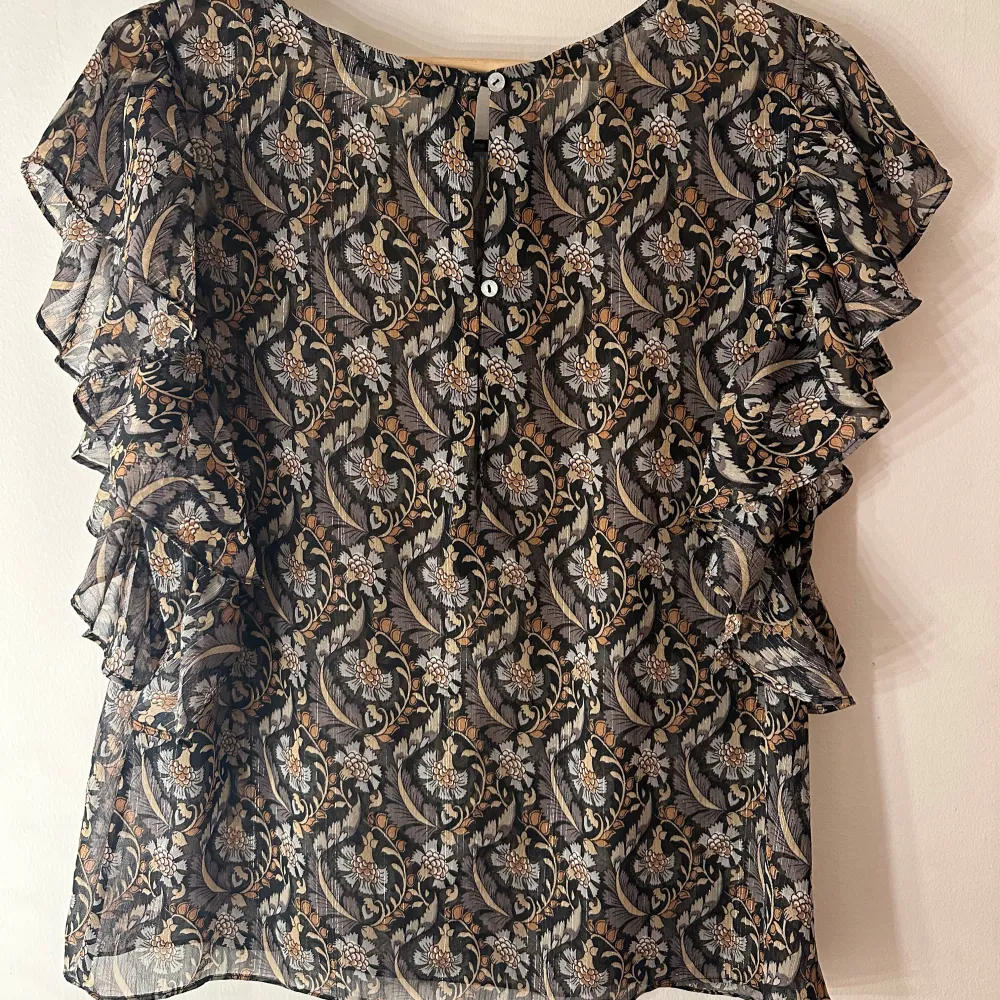 Snygg och elegant blus från Rue de Femme med ett vackert blommigt mönster i svart, beige och grå toner. Blusen har en avslappnad passform med volangdetaljer vid ärmarna som ger den en romantisk touch. Rund halsringning och knappar i ryggen. Perfekt för både vardag och fest. Något genomskinlig så kan behövas ett linne under.. Puserot.