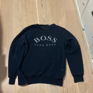 Mörkblå tröja från Hugo Boss - Säljer en stilren mörkblå tröja från Hugo Boss med logotypen tryckt på framsidan. Tröjan har långa ärmar och en rund halsringning. Perfekt för en avslappnad och snygg look.