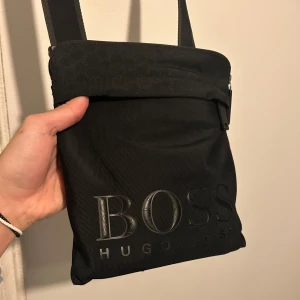 Svart axelväska från Hugo Boss - Snygg svart axelväska från Hugo Boss med justerbar rem och dragkedja. Limited edition från 2022. Perfekt för att bära med sig det nödvändigaste. Funkar till allt. Nypris ca 1200kr. Skick: 9/10. Mer bilder och beskrivning finns om så önskas. Pris kan diskuteras:) Mvh/ Philip