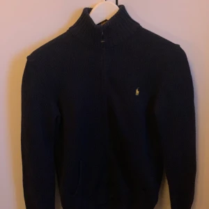 Mörkblå stickad tröja från Ralph Lauren - Säljer en snygg mörkblå stickad tröja från Ralph Lauren med dragkedja och hög krage. Tröjan har ett diskret broderat märke på bröstet och ribbade muddar vid ärmslut och nederkant. Perfekt för kyliga dagar!  Litet slitage i fickan går enkelt att laga. Köpt för 1250kr