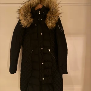 Svart dunjacka med pälsdetaljer - Snygg svart lång dunjacka med pälsdetaljer på huvan. Jackan har dragkedja och knappar framtill samt två fickor med dragkedja. Perfekt för kalla dagar.