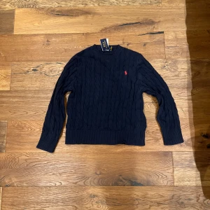 Mörkblå stickad tröja från Polo Ralph Lauren - Säljer en snygg mörkblå stickad tröja från Polo Ralph Lauren med kabelstickat mönster. Tröjan har en klassisk design med långa ärmar och ribbade kanter. Perfekt för kyligare dagar!