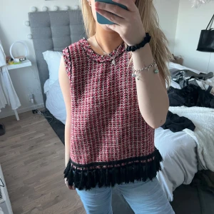 Röd och svart ärmlös topp med fransar - Snygg ärmlös topp i rött och svart med ett unikt mönster. Toppen har fransdetaljer längst ner som ger en trendig touch. Perfekt för att piffa upp din outfit!