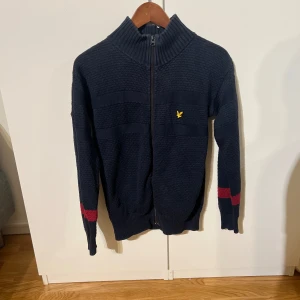 Mörkblå stickad tröja från Lyle & Scott - Säljer en mörkblå stickad tröja från Lyle & Scott med dragkedja och ribbad krage. Tröjan har långa ärmar med röda detaljer vid ärmsluten och ett litet gult emblem på bröstet. Perfekt för kyliga dagar!