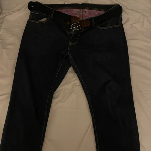 Mörkblå jeans från Oscar Jacobson - Snygga mörkblå jeans från Oscar Jacobson med en unik rosa mönstrad insida. Byxorna har en klassisk design med knappar och bälte. Perfekta för en stilren look. Byxorna är i storlek 34 men små i storleken så skulle nog säga att dem är i 30-31. Längden på dem är L30. 