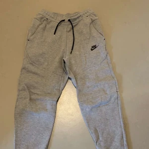 Grå mjukisbyxor från Nike tech - Säljer ett par grå mjukisbyxor från Nike med svart logga på sidan. Byxorna har snörning i midjan och sidofickor. Perfekta för en avslappnad stil.