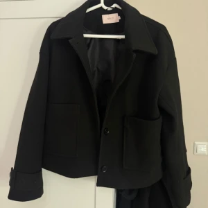 Svart kappa från Nelly - Snygg svart kappa från Nelly i storlek S. Jackan har en klassisk krage och knappar framtill. Den har långa ärmar med knappdetaljer vid ärmsluten och praktiska fickor framtill. Perfekt för en stilren look.