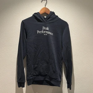 Svart hoodie från Peak Performance - Säljer en stilren svart hoodie från Peak Performance med broderad logga på bröstet. Tröjan har en klassisk design med känguruficka och ribbade muddar vid ärmslut och nederkant. Perfekt för en avslappnad look.
