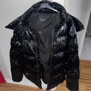 Svart glansig pufferjacka från Trapstar - Snygg svart pufferjacka från Trapstar med glansig finish och broderad logga på ryggen. Jackan har en hög krage och dragkedja framtill. Perfekt för kyliga dagar.