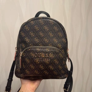 Mönstrad ryggsäck från Guess - Snygg liten  ryggsäck från Guess med brunt och guld mönster . Väskan har ett fack där fram och ett inner fack i väskan  och justerbara axelremmar. Kvitto och dustbag finns med. 