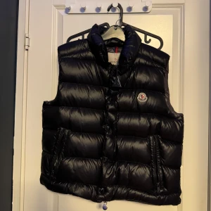Svart dunväst från Moncler - Säljer en stilren svart dunväst från Moncler med hög krage och tryckknappar. Västen har två praktiska fickor framtill och Monclers logotyp på bröstet. Perfekt för kyligare dagar.
