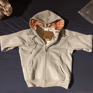 Grå huvtröja från Burberry - Snygg grå huvtröja från Burberry med dragkedja och klassiskt rutigt foder i huvan. Tröjan har långa ärmar och praktiska fickor framtill. Perfekt för en stilren och bekväm look.