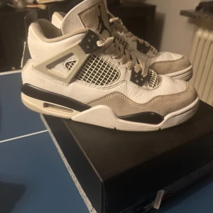 Air Jordan 4 Retro (GS) i vit och grå - Snygga Air Jordan 4 Retro sneakers i vit och neutral grå med svarta detaljer. Skorna har den klassiska Jumpman-loggan på tungan och hälen, samt ett nätmönster på sidorna. Perfekta för dig som älskar stil och komfort.