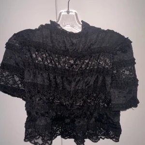 Svart spetsblus - Elegant svart spetsblus med korta ärmar och volangdetaljer. Perfekt för en stilfull look med en touch av romantik.