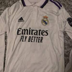 Säljer en vit långärmad Real Madrid-tröja från Adidas med Benzema och nummer 9 på ryggen. Tröjan har klubbens emblem och sponsorlogga på framsidan. Den har lila detaljer på kragen och ärmarna. Perfekt för fans av laget eller Benzema!