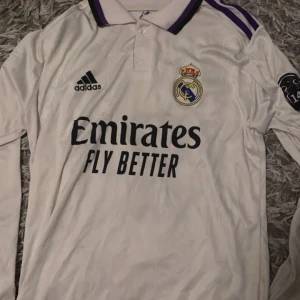 Vit långärmad Real Madrid-tröja - Säljer en vit långärmad Real Madrid-tröja från Adidas med Benzema och nummer 9 på ryggen. Tröjan har klubbens emblem och sponsorlogga på framsidan. Den har lila detaljer på kragen och ärmarna. Perfekt för fans av laget eller Benzema!