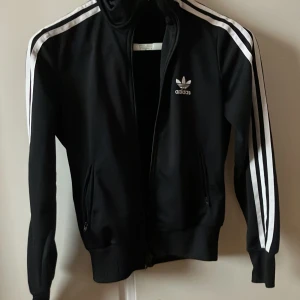 Svart träningsjacka från Adidas - Säljer en klassisk svart Adidas träningsjacka med vita ränder längs ärmarna och det ikoniska trefoil-loggan på bröstet och ryggen. Jackan har dragkedja och ribbade muddar. Perfekt för både träning och vardag!