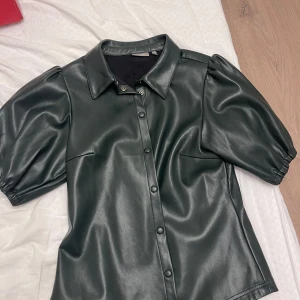 Svart skinnskjorta med puffärmar - Snygg svart skinnskjorta med korta puffärmar och knappar framtill. Perfekt för en cool och edgy look. Passar bra till både vardag och fest. Skjortan har en klassisk krage och är i mycket bra skick.