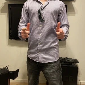 Lila rutig skjorta från Eton - Säljer en stilren lila rutig skjorta från Eton i storlek 38. Skjortan har en slim passform och är långärmad, perfekt för både kontoret och fest. Materialet känns lyxigt och bekvämt. Den har klassiska knappar framtill och en snygg krage. Passar perfekt till både jeans och kostymbyxor!