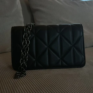 Svart quiltad axelväska med kedja - Snygg svart axelväska i quiltat mönster med en cool kedja som axelrem. Väskan har en praktisk storlek och stängs med en tryckknapp. Insidan har flera fack. Jättefint skick då den inte kommit till användning på länge. Tvivla inte på att höra av dig om du funderar på något !